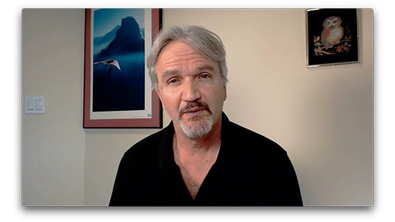 A man's face is visible on a computer screen, accompanied by another screen in a modern workspace.